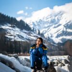 woman in blue jacket and blue backpack sitting on rock near snow covered mountain during daytime